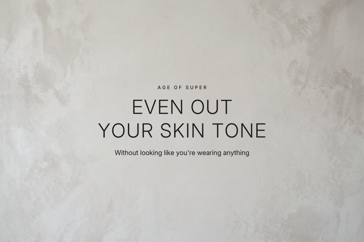 Centered text "Even out your skin tone" with the line "Without looking like you're wearing anything" on a smooth warm gray stone surface that is mottled at the edges and even in the center